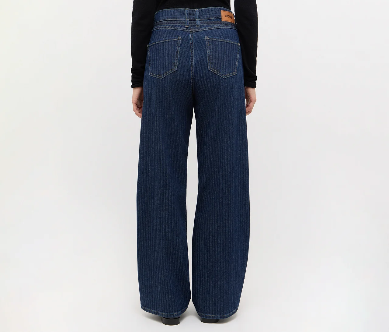 Rückansicht einer Person in dunkelblauen, weit geschnittenen Jeans mit feinen vertikalen Streifen und einem schwarzen Langarmshirt. Die Jeans hat Gesäßtaschen und ein braunes Etikett.