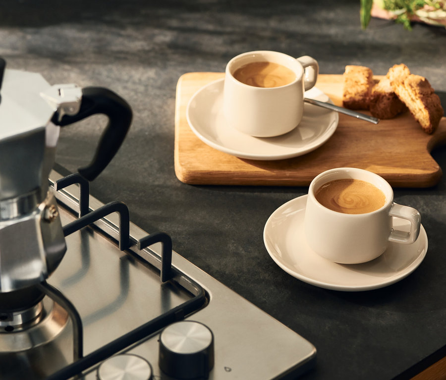 Silberne Mokkakanne auf einem Gasherd neben zwei hellen Kaffeetassen mit Untertassen und Biscotti auf einem Holzbrett.