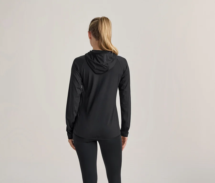 Frau von hinten, trägt eine schwarze Kapuzenjacke und schwarze Leggings. Ihr blondes Haar ist zu einem Pferdeschwanz gebunden.