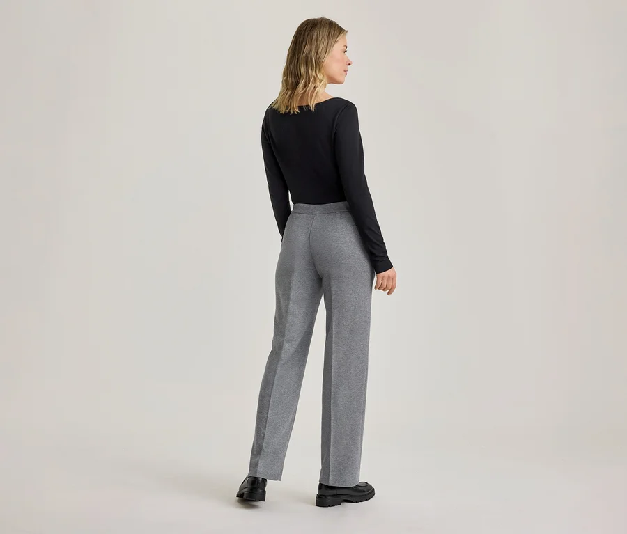 Eine Frau von hinten, die ein schwarzes Langarmshirt, eine graue weite Hose und schwarze Schuhe trägt. Sie steht mit leicht nach rechts gedrehtem Kopf.