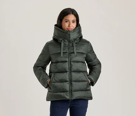 Frau in dunkelgrüner Steppjacke mit hohem Kragen und Kapuze, Hände in den Taschen, dazu dunkle Jeans.