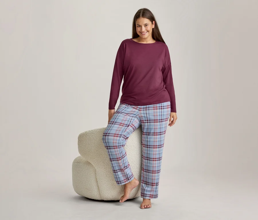 Lächelnde Frau in weinrotem Langarmshirt und hellblau, rot und weiß karierter Pyjamahose, die mit einem Fuß auf einem flauschigen weißen Hocker steht.