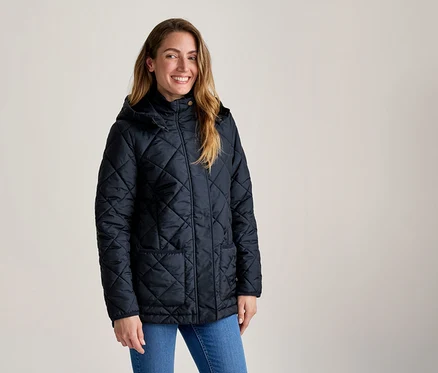 Lachende Frau mit langen Haaren trägt eine dunkelblaue Steppjacke mit Kapuze und blaue Jeans.