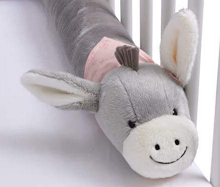 Grauer Plüsch-Esel als Bettumrandung mit weißer Schnauze und rosa Stoff um den Hals, liegt auf einem weißen Laken im Gitterbett.