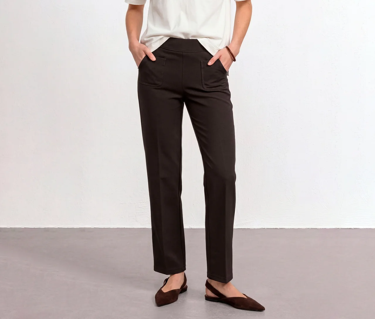Eine Person trägt eine braune Street One Stoffhose, die Hände in den Taschen, ein weißes T-Shirt und braune flache Schuhe.