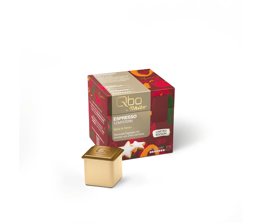 Eine rote und goldene Packung Qbo Espresso Zimtstern Kapseln, als Limited Edition mit Zimt- und Nussaromen, steht neben einer einzelnen goldenen Kapsel und sternförmigen Zimtsternen.