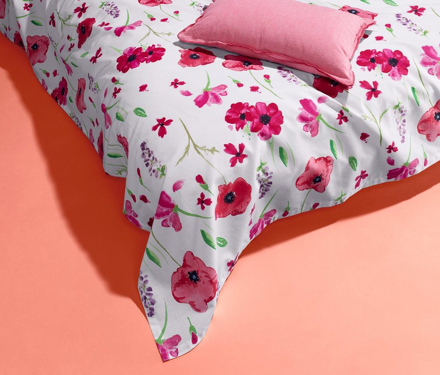 Eine weiße Renforcé-Multifunktions-Überwurf mit Blumenmuster und ein rosa Kissen auf korallefarbenem Hintergrund.