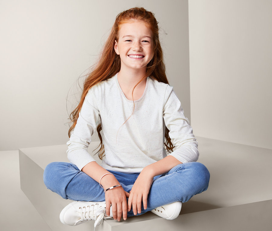 Mädchen sitzt im Schneidersitz und lächelt, trägt 2 Langarmshirts.