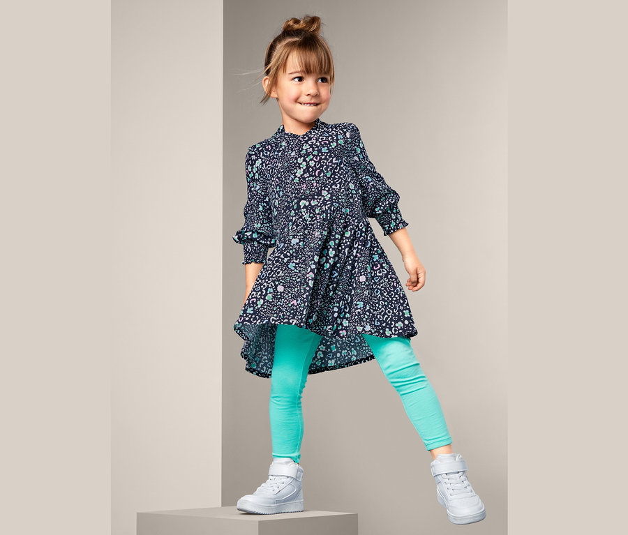 Mädchen steht auf einem grauen Würfel und trägt ein Webkleid, grüne Leggings und weiße Schuhe.
