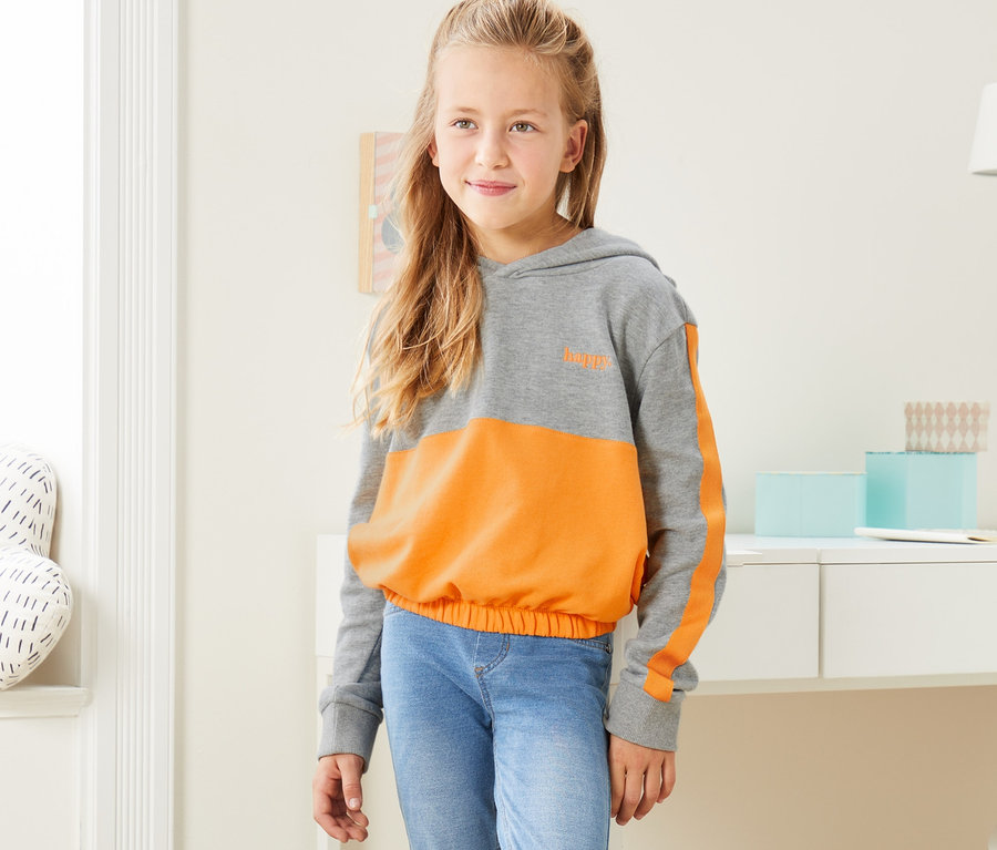 Mädchen steht und trägt einen grau-gelb melierten Cropped-Hoodie, Jeans und Turnschuhe.