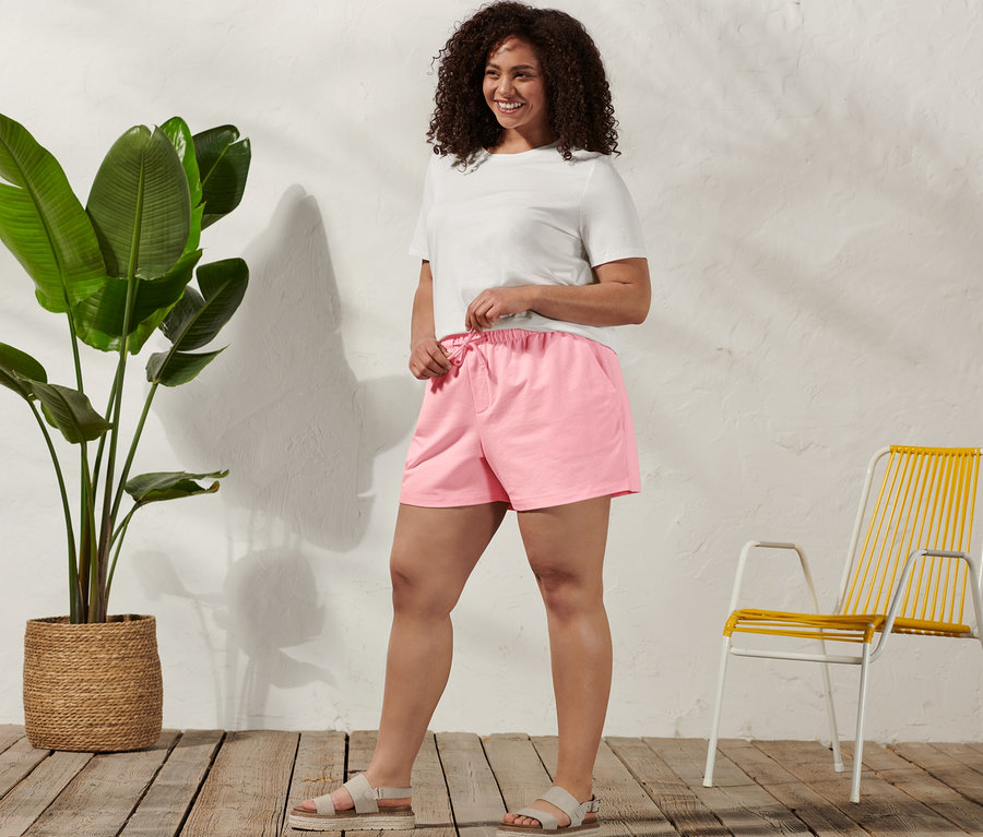 Eine Frau steht und zieht die Kordel ihrer Sweatshorts fest, sie trägt ein Basic T-Shirt, weiß, und Komfortsandale.