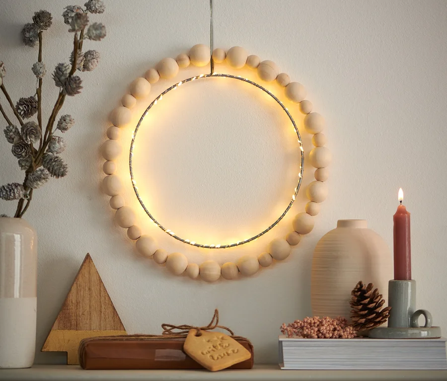 Ein LED-Ring mit Holzperlen hängt an der Wand über einem Kaminsims mit Dekorationen.