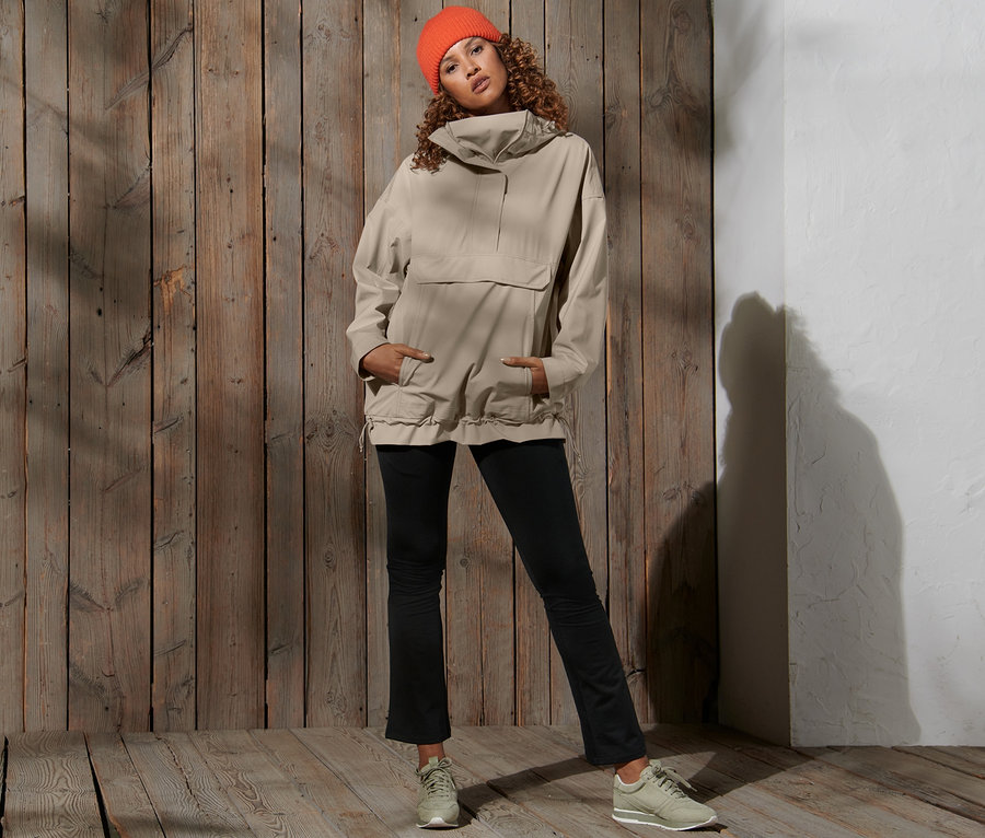 Frau posiert vor einem Holzhintergrund und trägt eine orangefarbene Mütze und eine beige Oversized-Funktionsregenjacke.