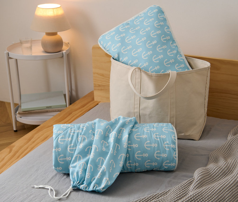 Ein Reise-Bettenset mit Ankermotiv in einer beigen Tasche und zusammengerollt auf einem Bett.