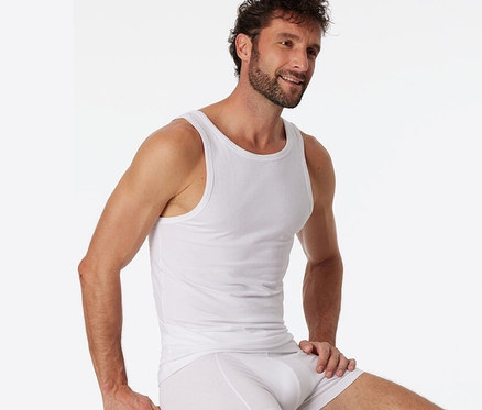 Mann sitzt auf einem Hocker und trägt 2 SCHIESSER Unterhemden mit Bio-Baumwolle, weiß.