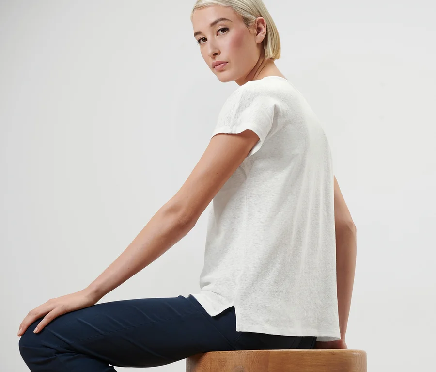 Frau sitzt auf einem Holzhocker und trägt ein cremeweißes Leinen-Shirt.