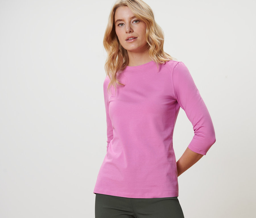 Blonde Frau posiert in einem rosa Shirt mit 3/4-Arm.