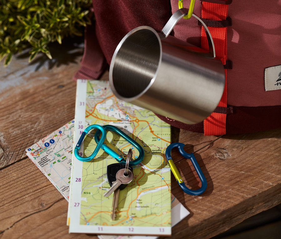 Eine Edelstahl Tasse hängt mit einem der 4 Karabinerhaken an einem Rucksack. Auf einer Karte liegen Schlüssel mit einem weiteren Karabiner.