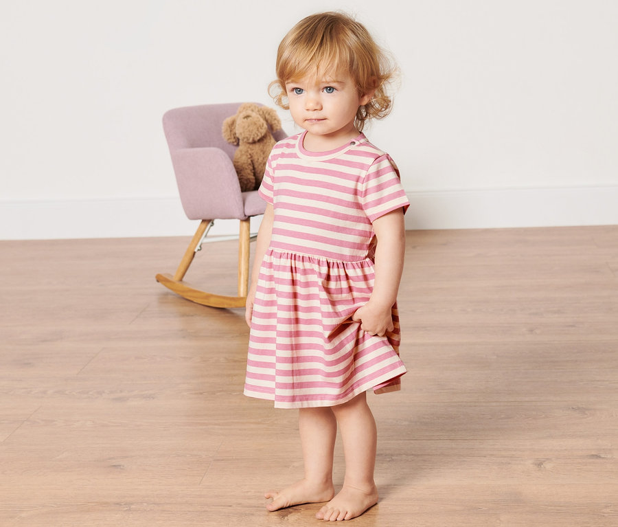Mädchen steht auf dem Boden und trägt ein Baby-Jerseykleid mit rosa Streifen.