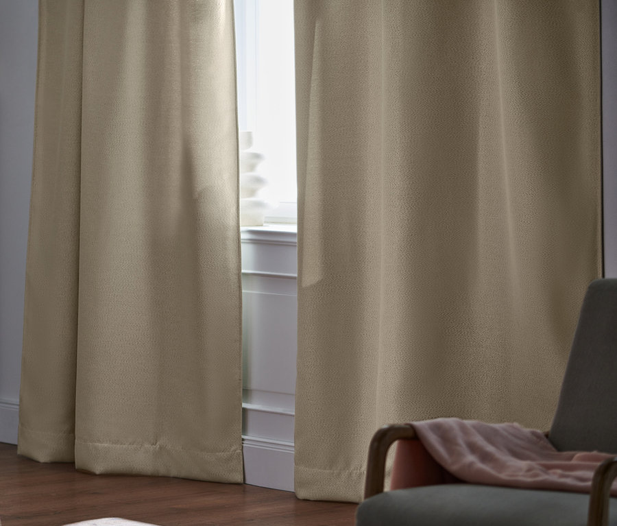 Schlafzimmer mit beigen 2 Verdunkelungs-Vorhangschals am Fenster, Bett und grauem Sessel.