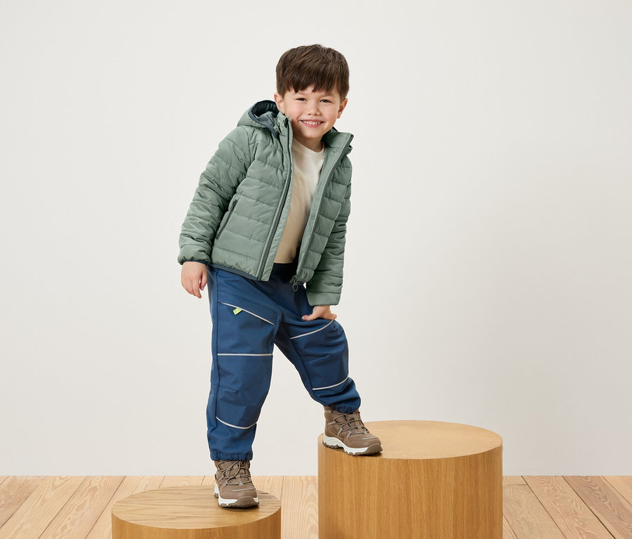 Lächelnder Junge posiert auf einem Holzsockel in grüner Jacke, blauer Hose und braunen Schuhen.