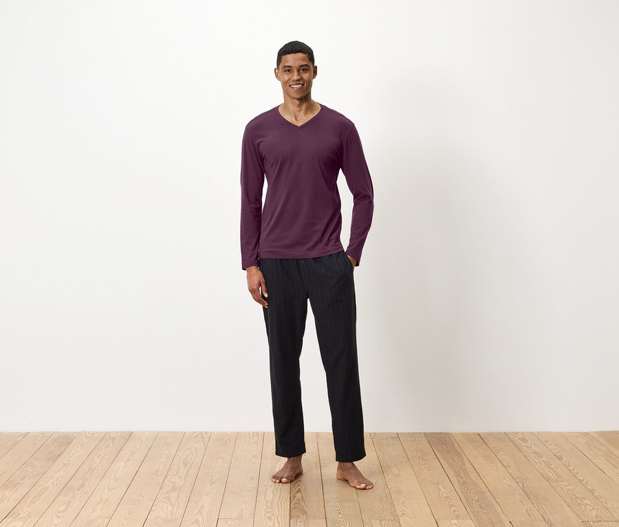 Ein Mann steht barfuß auf einem Holzboden und trägt ein weinrotes Langarmshirt und eine schwarze Hose.