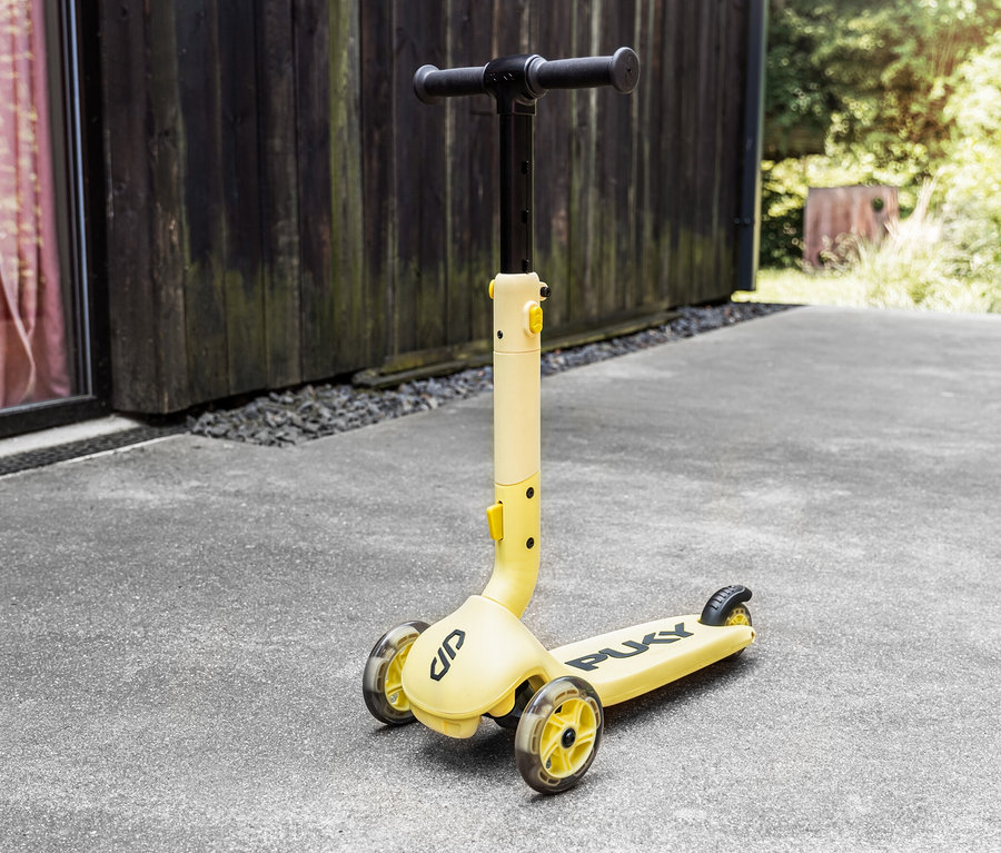 Ein gelber PUKY Roller PUSH steht auf einem Betonboden vor einem Holzgebäude.