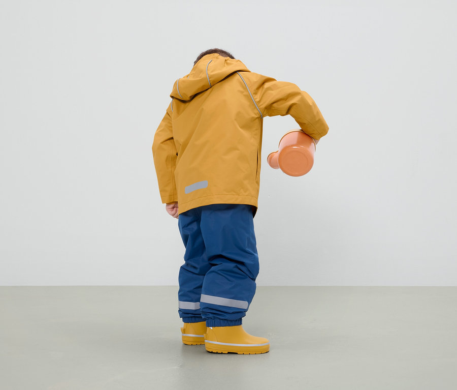 Ein Junge in einer senffarbenen Jacke, blauen Hosen und gelben Gummistiefeln hält eine orangefarbene Gießkanne.
