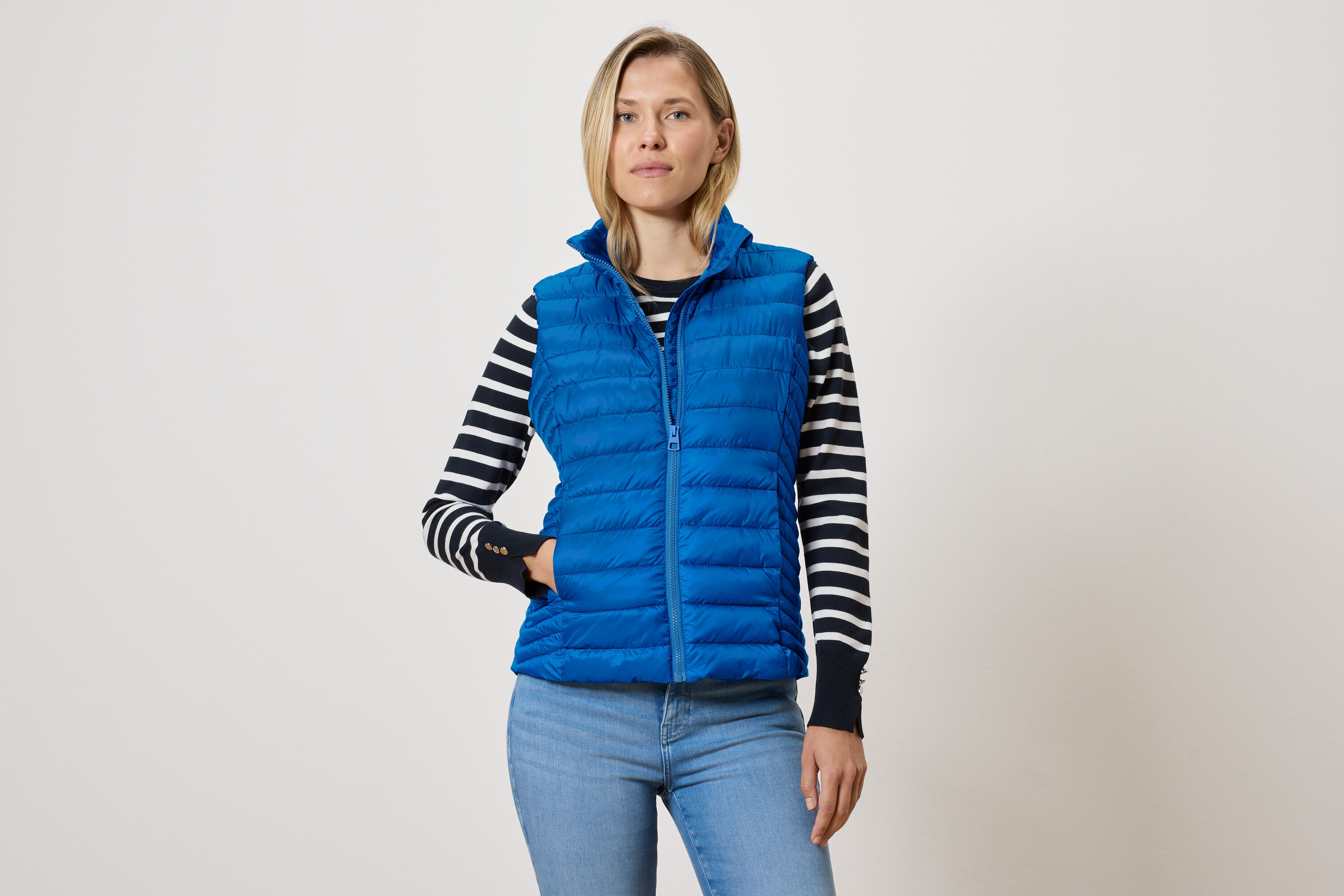Frau trägt eine blaue Steppweste mit Reißverschluss, einen schwarz-weiß gestreiften Pullover und blaue Jeans.