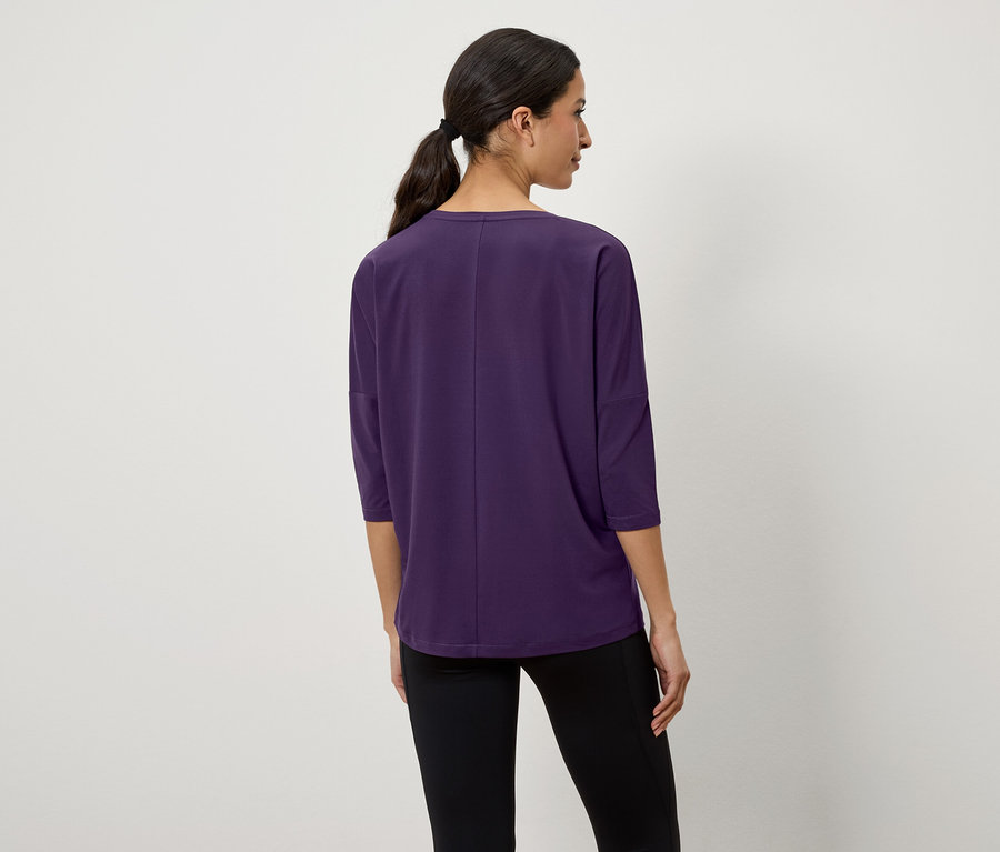 Frau mit dunklen Haaren, die zu einem Pferdeschwanz zusammengebunden sind, steht mit dem Rücken zur Kamera und trägt ein lila Dreiviertelarm-T-Shirt und schwarze Leggings.