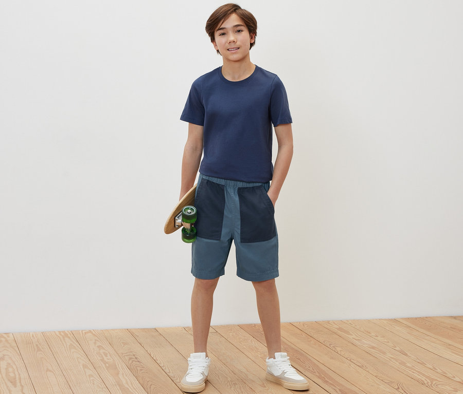 Junge hält ein Skateboard und trägt Kinder-Bermudashorts.