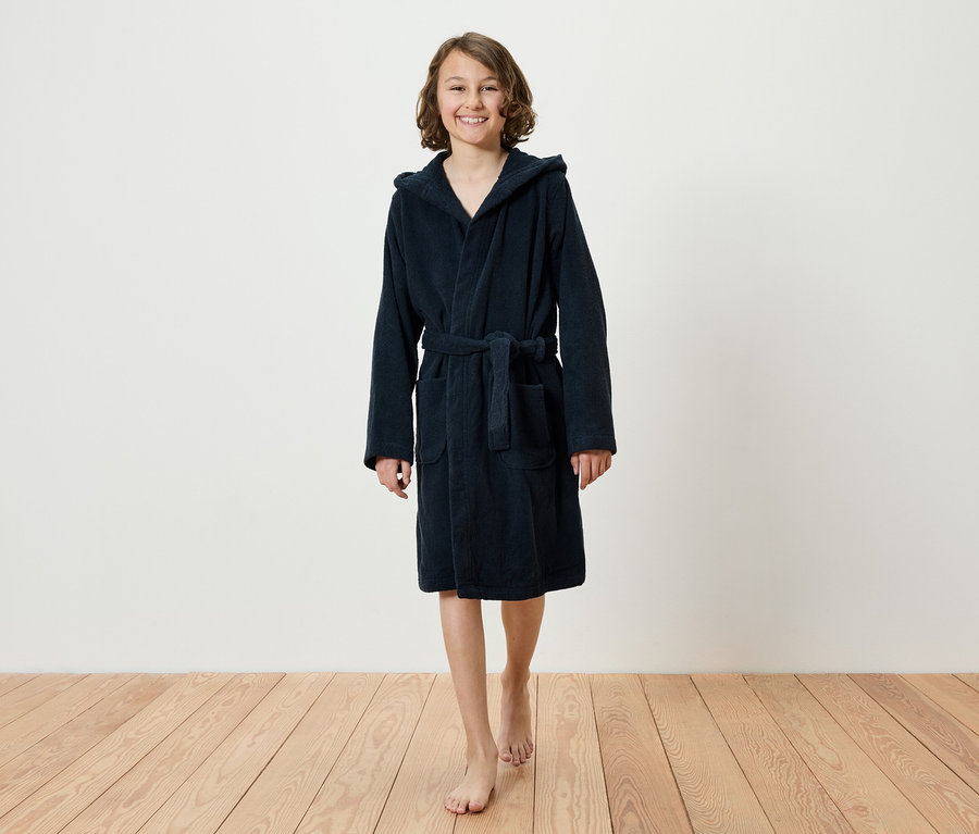 Lächelndes jugendliches Mädchen geht barfuß auf einem Holzboden und trägt einen dunkelblauen Bademantel mit Kapuze.
