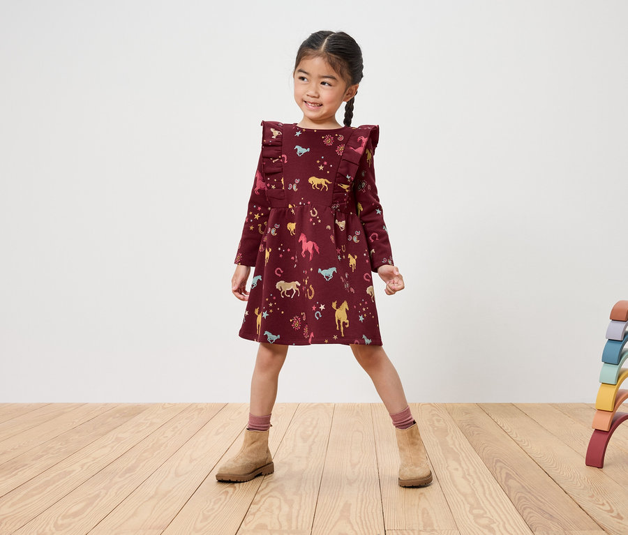 Lächelndes Mädchen in bordeauxfarbenem Kleid mit Pferdeaufdruck und braunen Stiefeln steht auf einem Holzboden neben einem regenbogenfarbenen Holzspielzeug.