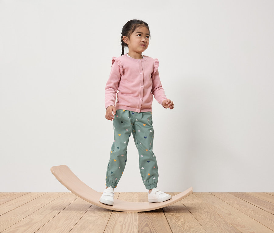 Mädchen steht auf einem hölzernen Balancebrett, trägt einen rosa Cardigan und grüne Hosen mit Herzen.
