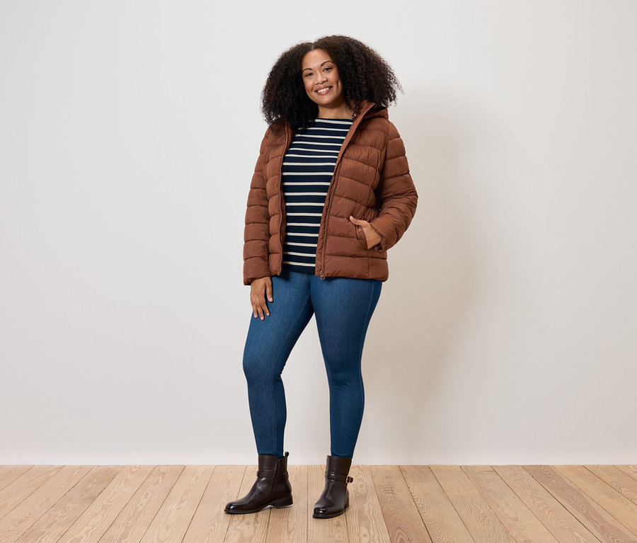 Frau mit lockigem Haar posiert in brauner Steppjacke, blauen Jeans und braunen Stiefeln.