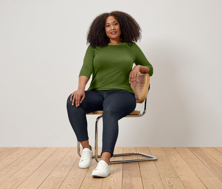 Eine Frau mit lockigem Haar sitzt auf einem Stuhl und trägt ein grünes Dreiviertelarm-Shirt, dunkle Jeans und weiße Turnschuhe.