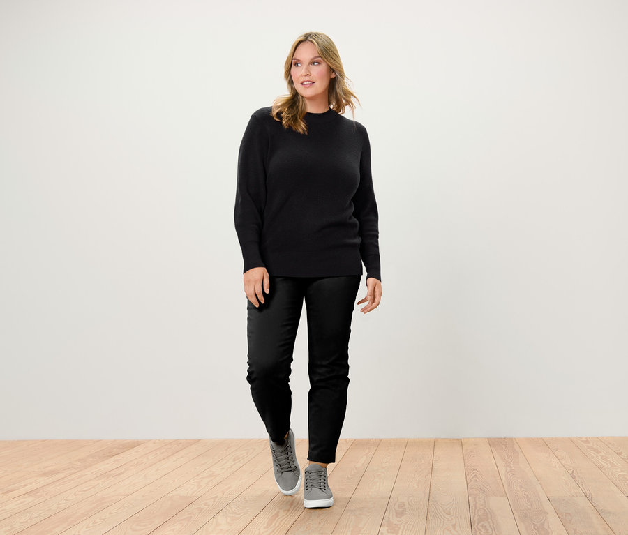 Frau geht auf Holzboden und trägt einen schwarzen Pullover, eine schwarze Hose und graue Turnschuhe.