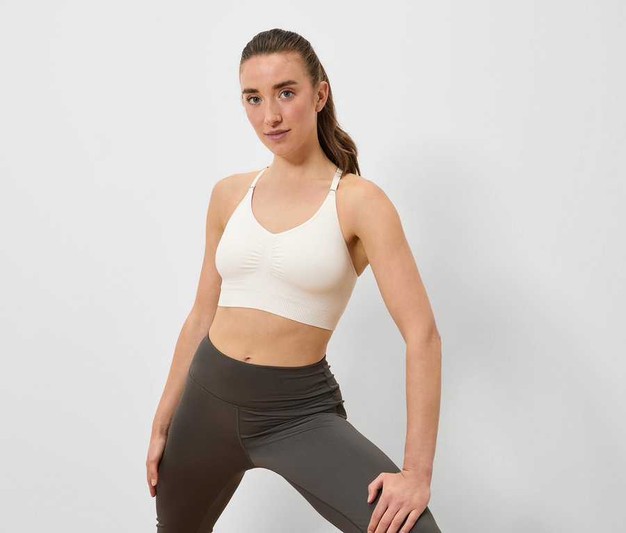 Frau posiert in einem cremefarbenen Sport-BH und grauen Leggings vor einem weißen Hintergrund.