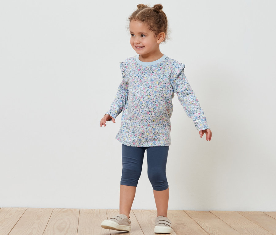 Ein kleines Mädchen posiert in 3/4-Leggings und einem geblümten Oberteil. Sichtbar ist eine der 5 Kinder-3/4-Leggings.