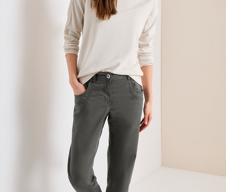 Lachende Frau mit langen braunen Haaren, trägt ein helles Langarmshirt, eine dunkelgraue CECIL Hose und silberne Sneaker.