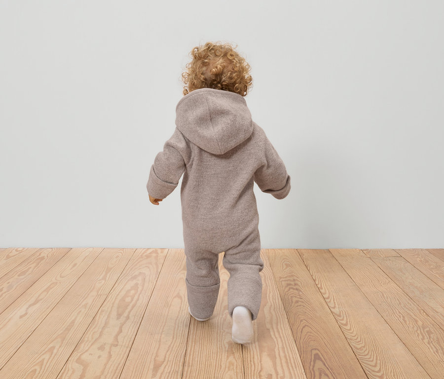 Rückansicht eines Kleinkindes mit lockigem Haar, das einen beigen Wollwalkoverall mit Kapuze und weiße Socken auf einem Holzboden trägt.