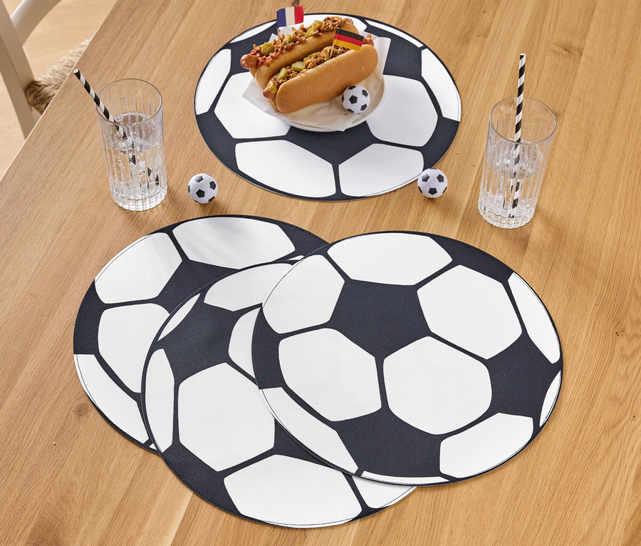 Zwei Hotdogs mit Flaggen auf einem Teller mit Fußballmuster, daneben Gläser mit Strohhalmen und Fußball-Dekoration auf einem Holztisch.