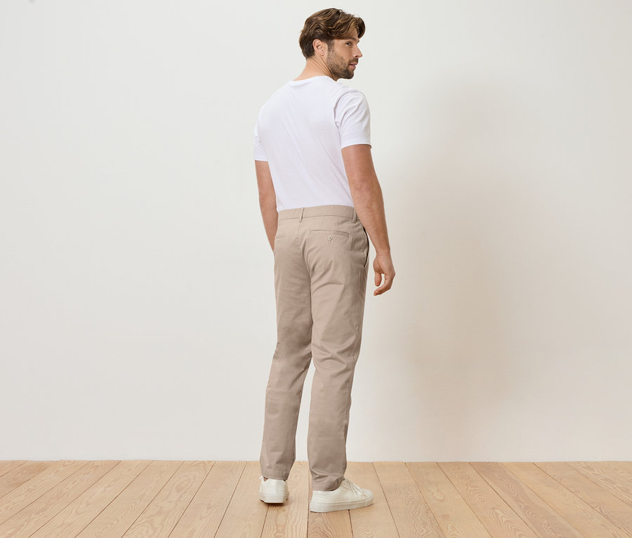 Mann von hinten in beigen Hosen und weißem T-Shirt, stehend auf Holzboden vor einer weißen Wand.