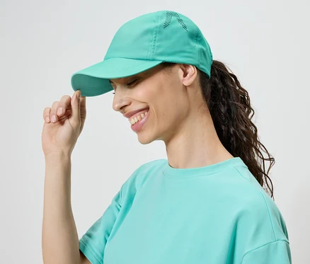 Lächelnde Frau mit einem türkisfarbenen Cap und T-Shirt, die den Schirm der Kappe berührt.