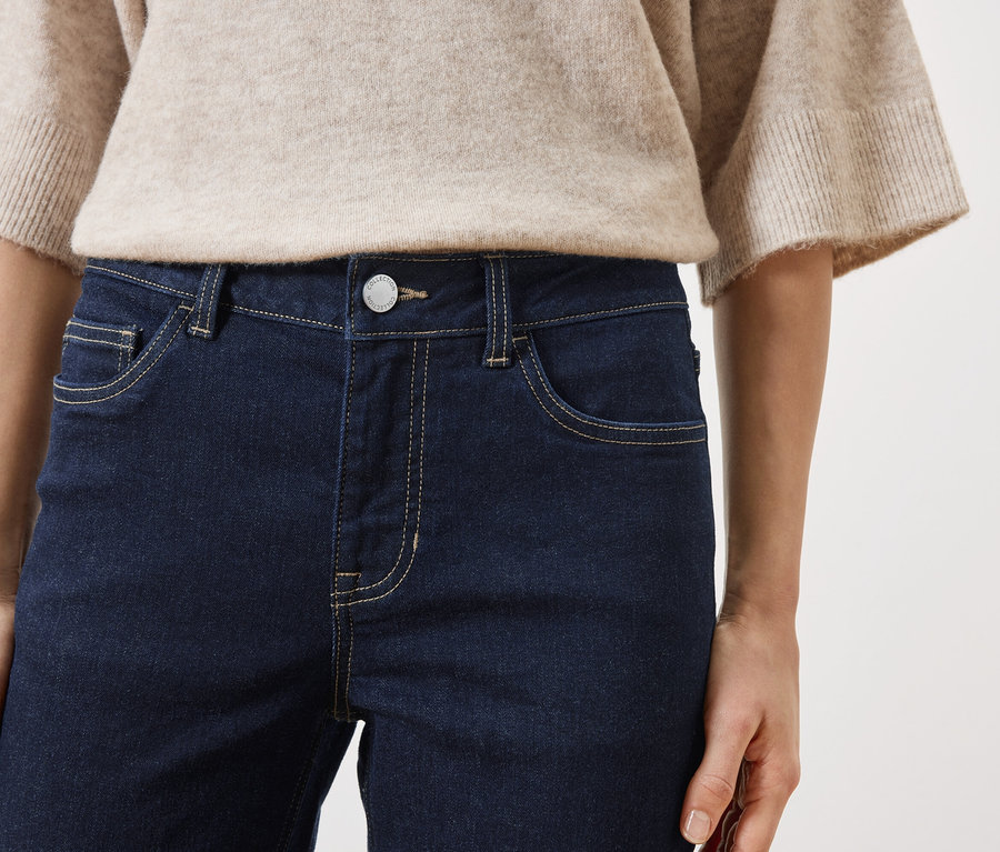 Nahaufnahme einer Frau in dunkelblauen Jeans und beigem Pullover vor einem weißen Hintergrund.