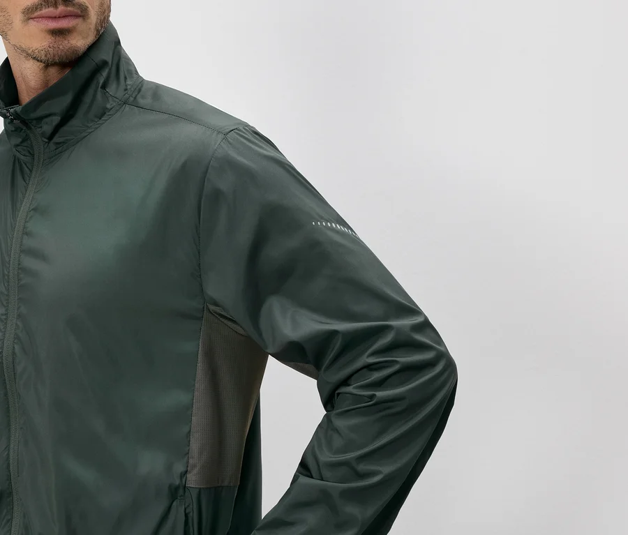 Mann in olivgrüner Jacke mit Reißverschluss und Stehkragen, Seitenpartien aus Mesh und reflektierenden Details am Ärmel.