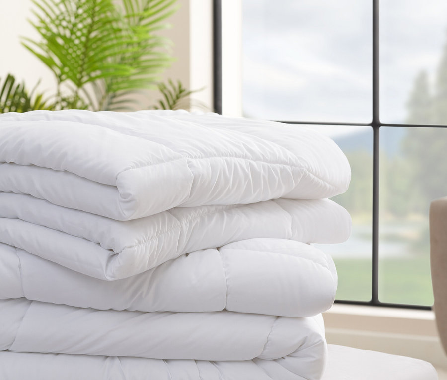 Ein Stapel weißer Bettdecken liegt auf einem Bett vor einem Fenster und einer Pflanze.