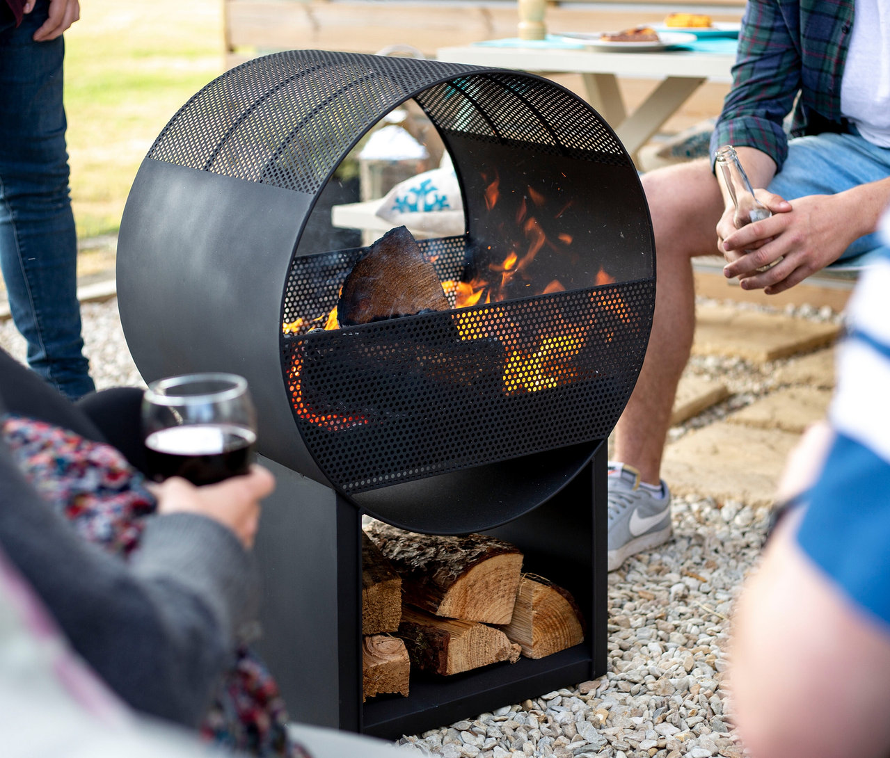 Personen sitzen um die Buschbeck Feuerstelle »Porthole«, in der Holz brennt. Eine Person hält ein Weinglas, eine andere eine Bierflasche.