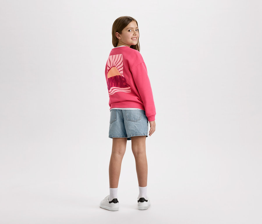 Mädchen von hinten, trägt einen pinken Pullover mit einem Sonnenaufgangsdesign, blaue Jeansshorts und weiße Turnschuhe.