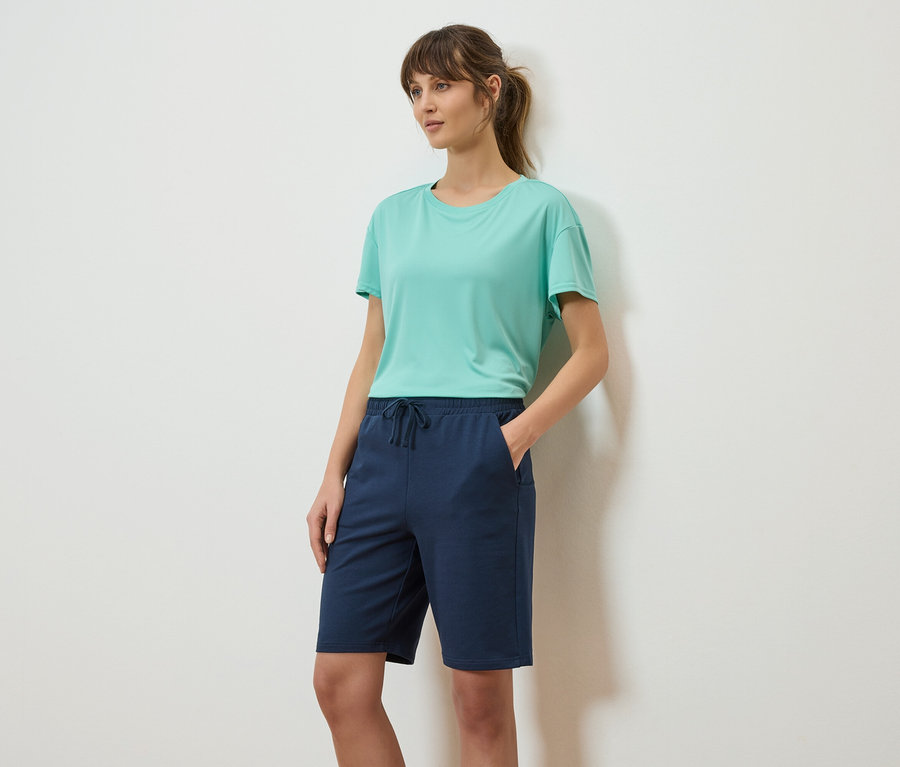 Frau mit Pferdeschwanz, trägt ein türkisfarbenes T-Shirt und marineblaue Shorts.